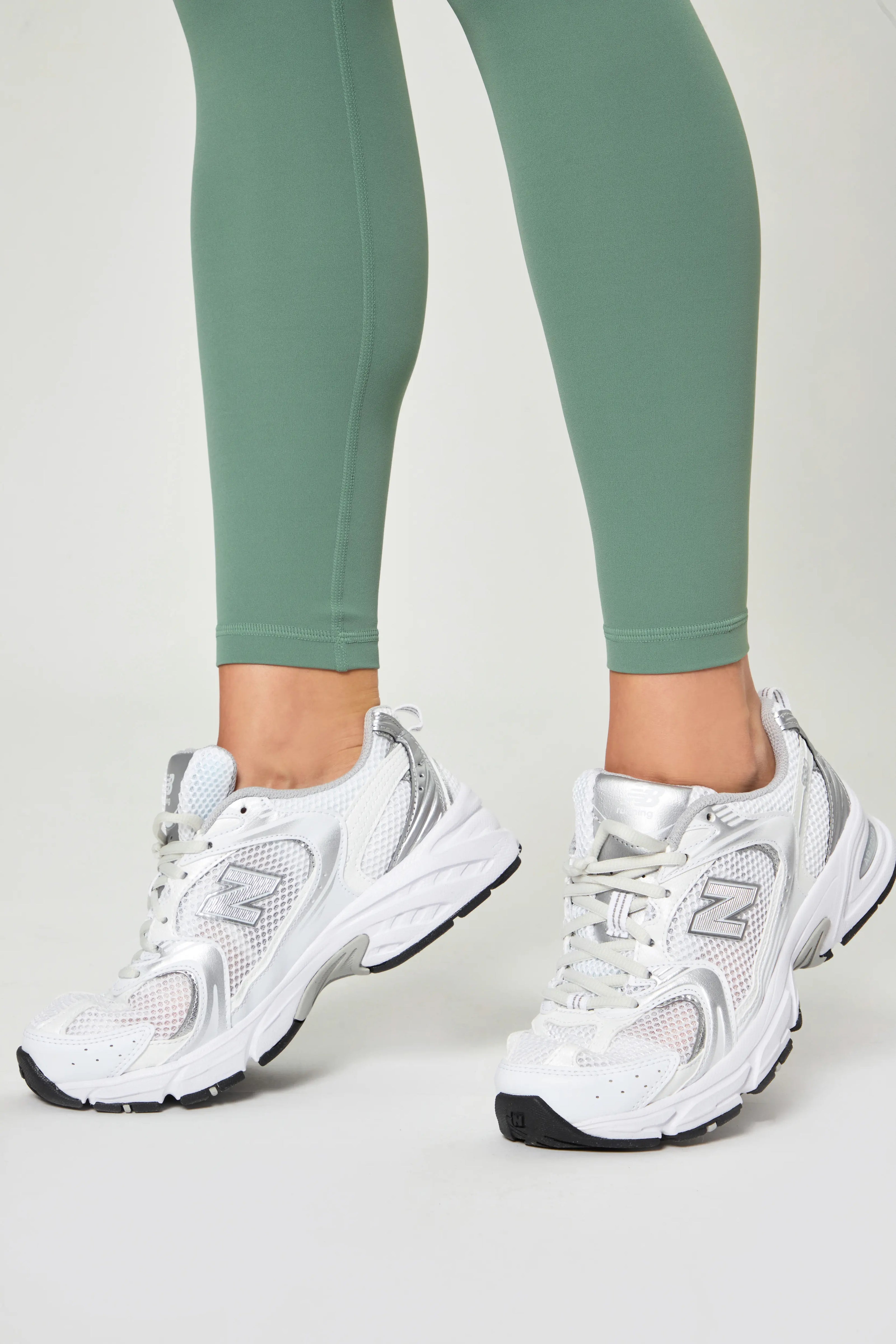 Close-up of legs wearing Базові легінси темно-зеленого кольору with white athletic sneakers on a light background